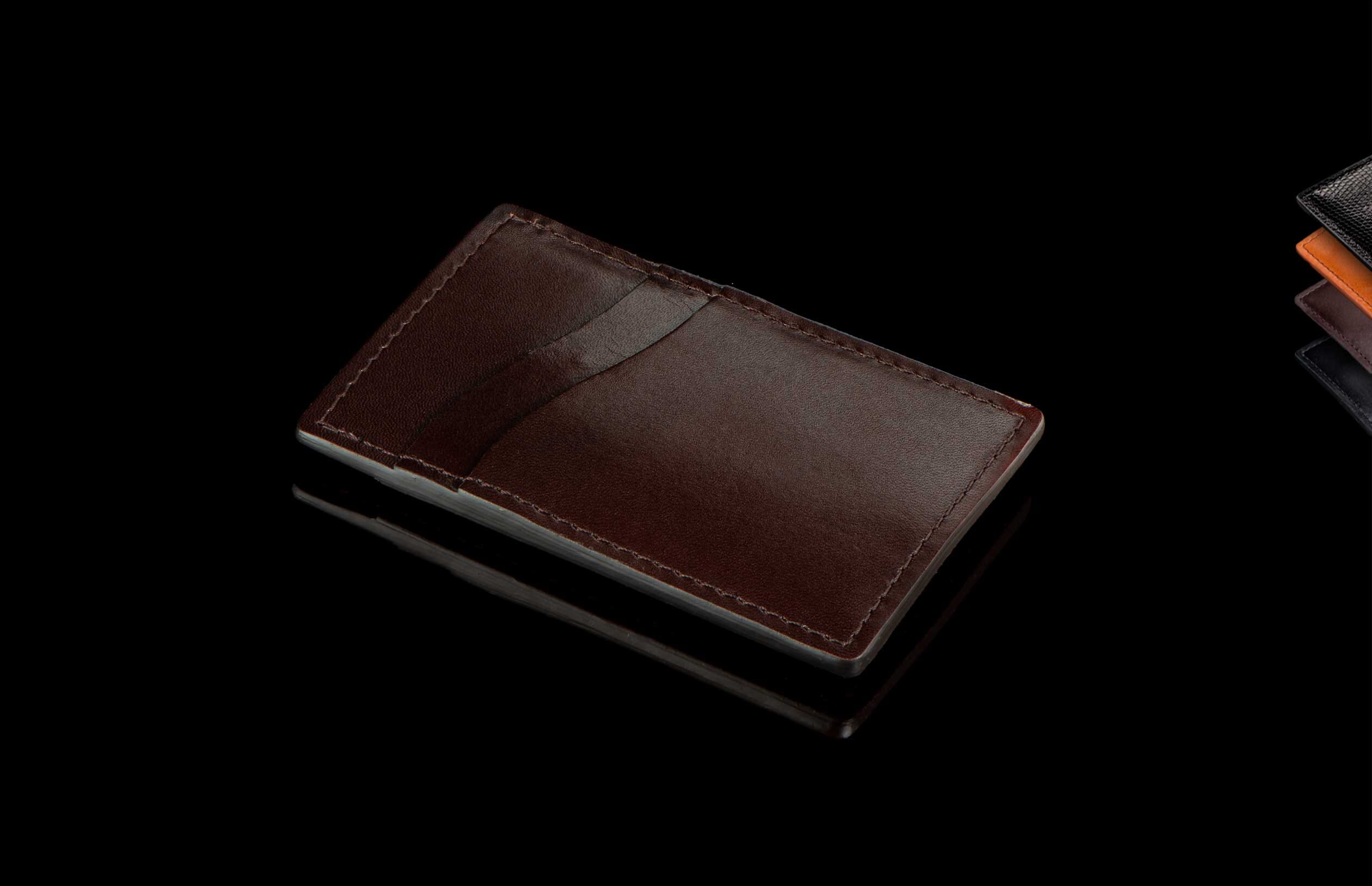 Dark brown leather Card & Cash Wallet with prominent stitching, reflecting on a glossy black surface. Other wallets are partially visible in the background.