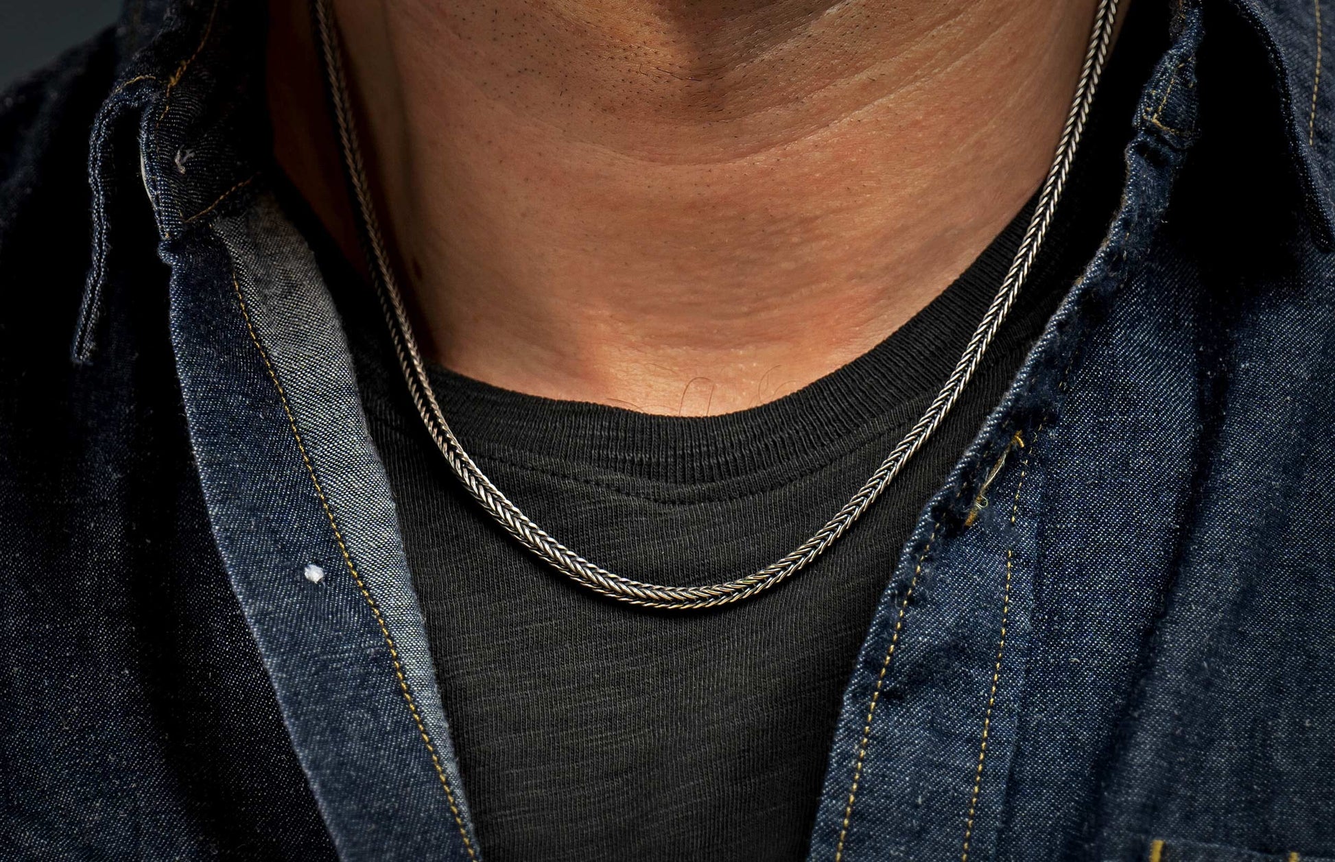 Silver-toned braided chain necklace worn on a man's neck over a dark t-shirt and denim shirt.