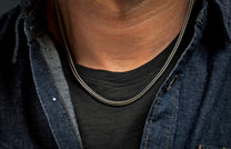 Silver-toned braided chain necklace worn on a man's neck over a dark t-shirt and denim shirt.