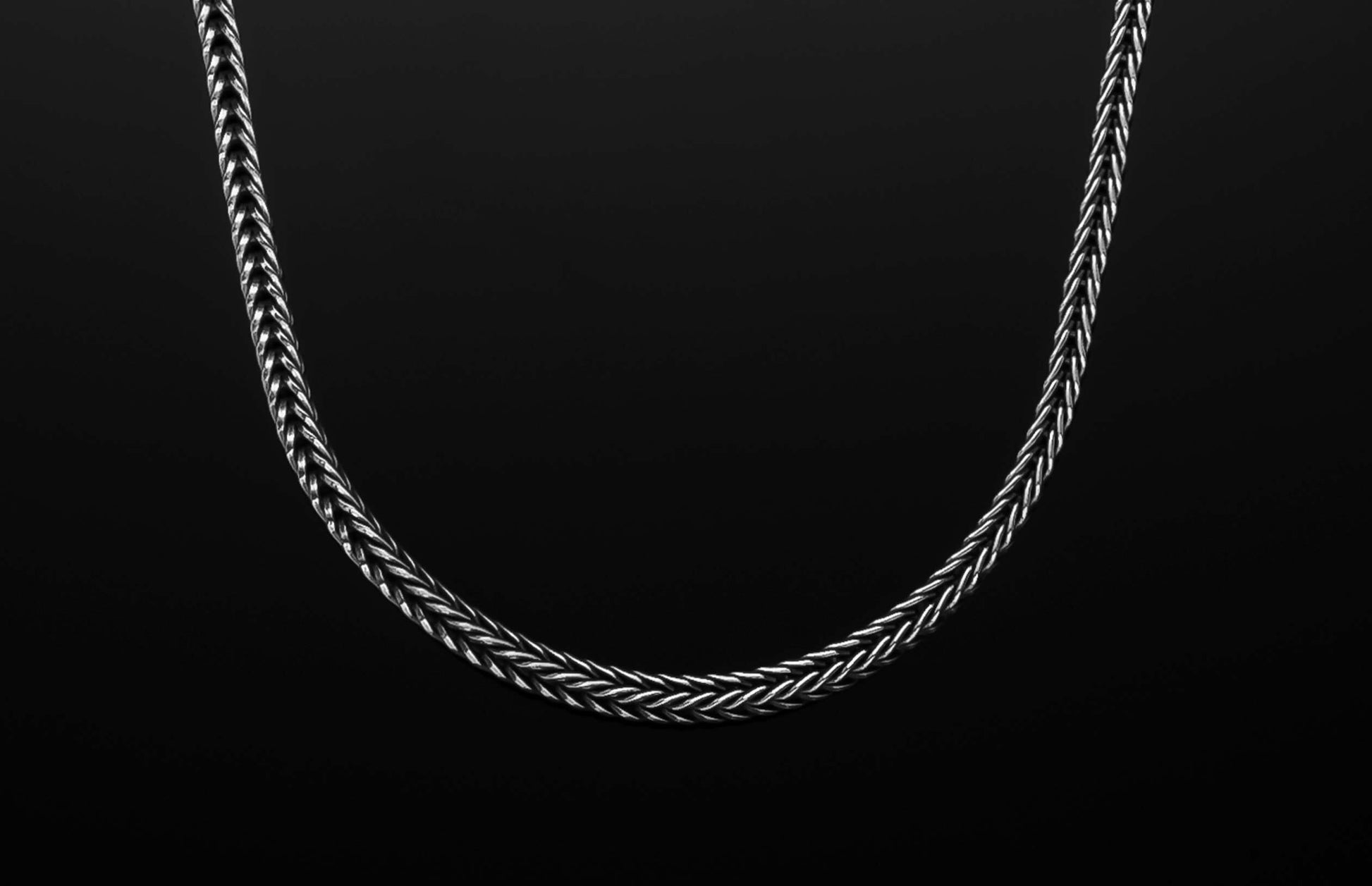 A silver braided chain necklace with intricately woven links, elegantly curved on a dark background.