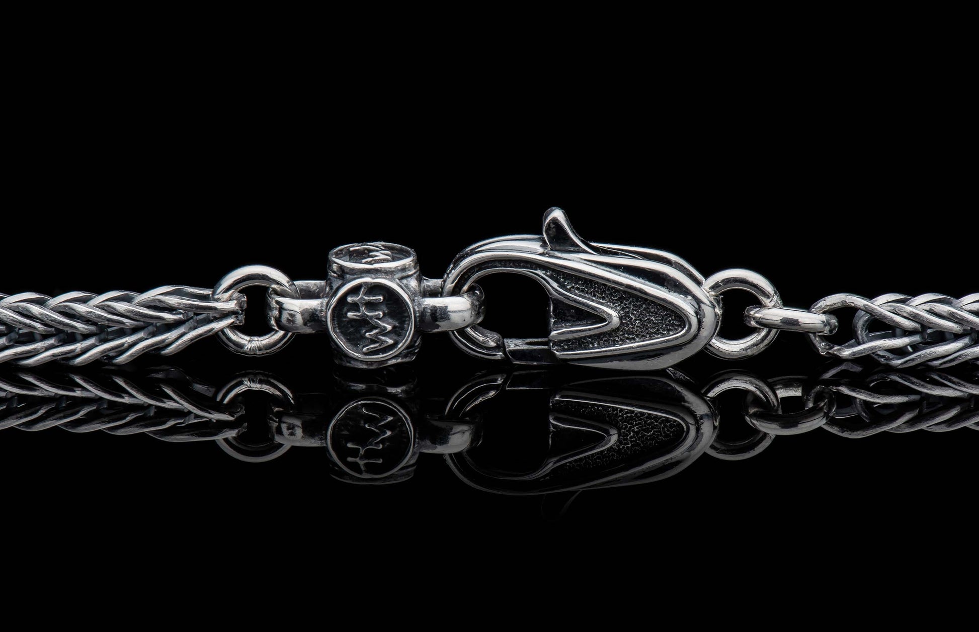 Silver braided chain with an ornate lobster clasp and 'HM' engraved barrel connector on a reflective surface.
