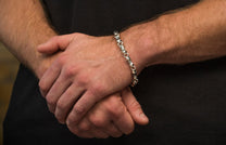 Chunky silver bracelet with detailed, decorative links worn on a man's wrist, hands clasped over a dark shirt.