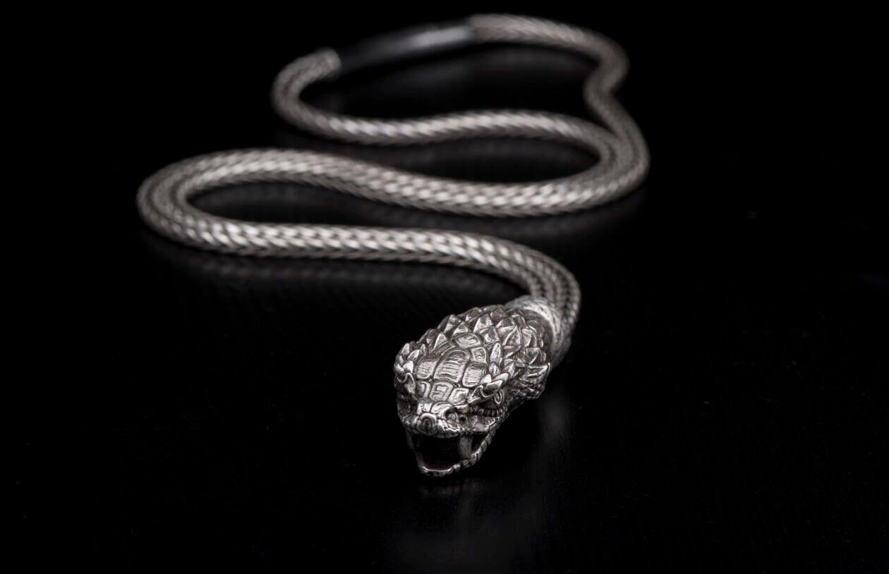 Silver snake-shaped jewelry with a braided body and detailed head with open jaws, on a black background.