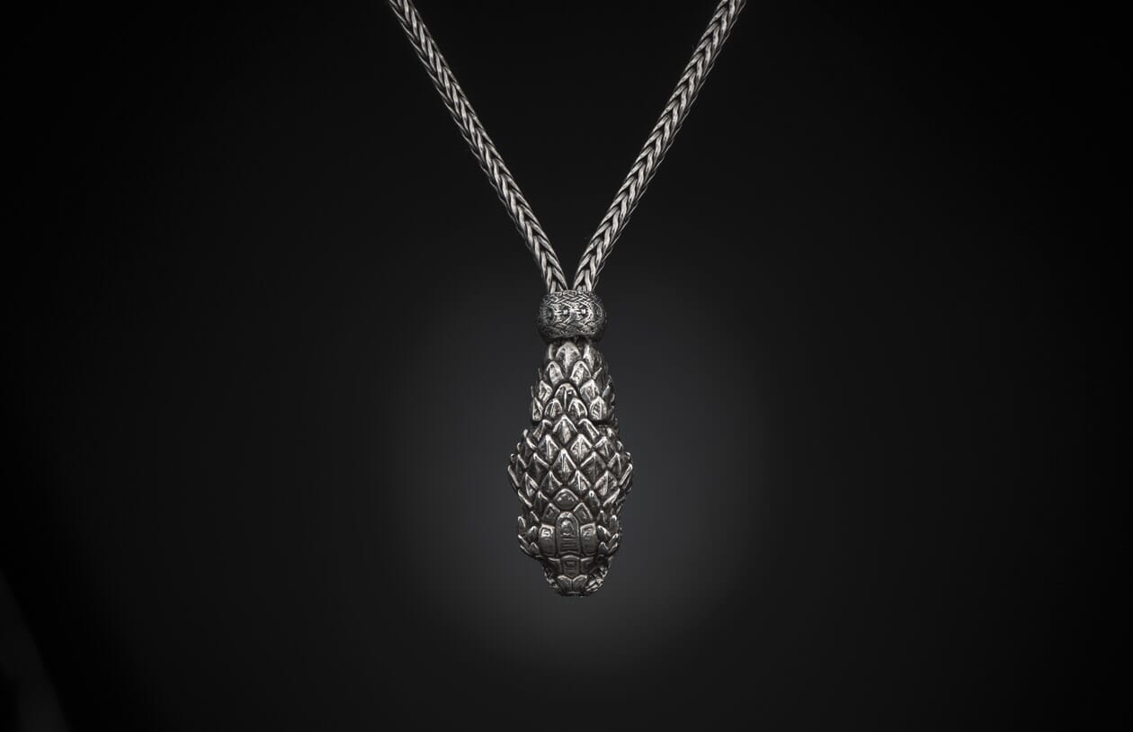 Silver braided necklace with a textured, tapered pendant featuring overlapping scale-like elements, against a black background.