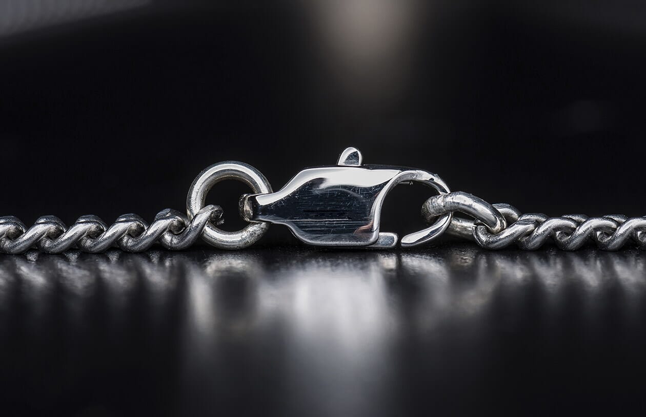 Silver chain necklace with a polished lobster clasp and textured links, viewed close-up on a dark reflective surface.