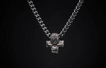 Silver metal necklace with a textured hammer-shaped pendant featuring a stylized face, centered on a black background.
