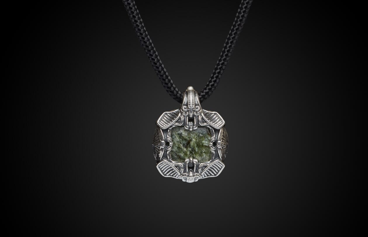 Silver-toned metal pendant with a dark green textured stone, featuring intricate designs, on a black braided cord.