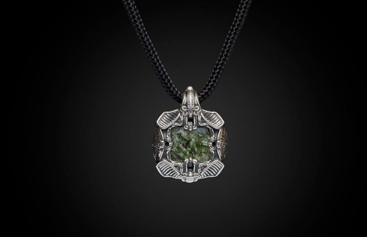 Silver-toned metal pendant with a dark green textured stone, featuring intricate designs, on a black braided cord.