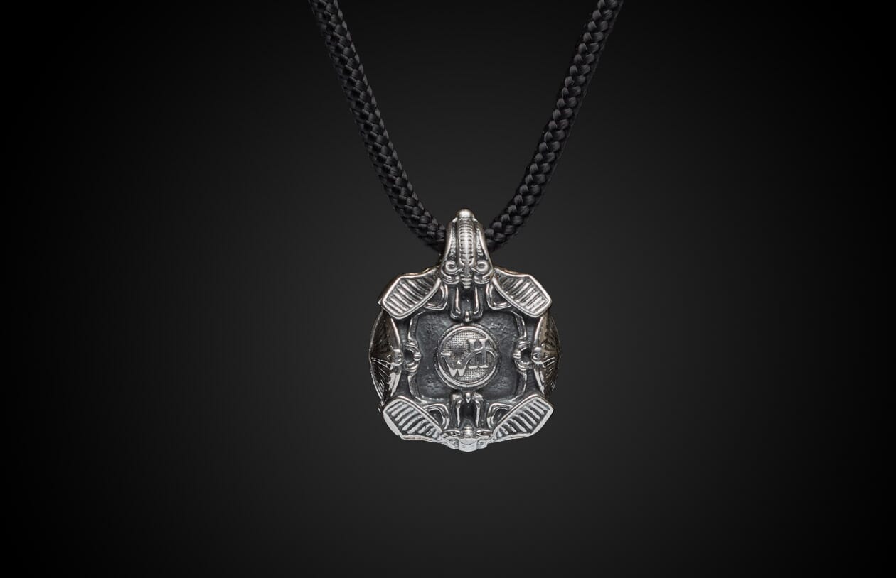 Silver Krypton pendant featuring intricate etched designs, a central circular emblem, and wing-like elements, on a black braided cord.