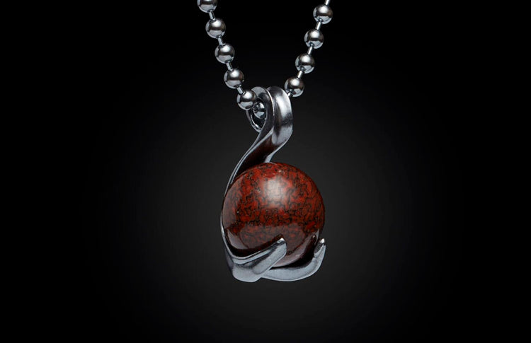 Silver-toned metal necklace pendant holding a spherical dark red stone with black speckled patterns, on a beaded chain.
