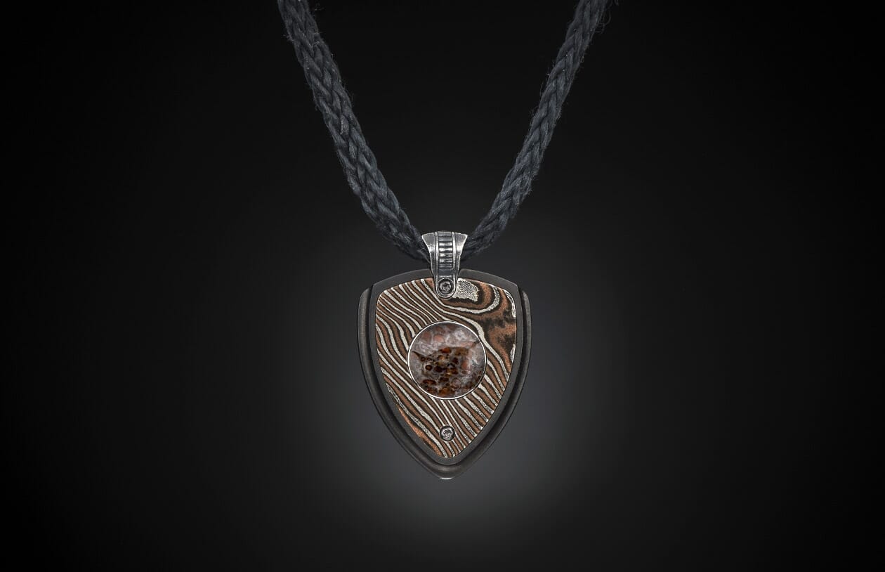 Pendant necklace with a shield-shaped pendant made of wavy patterned metal and a central reddish-brown stone, on a black braided cord.