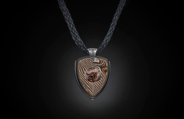 Pendant necklace with a shield-shaped pendant made of wavy patterned metal and a central reddish-brown stone, on a black braided cord.