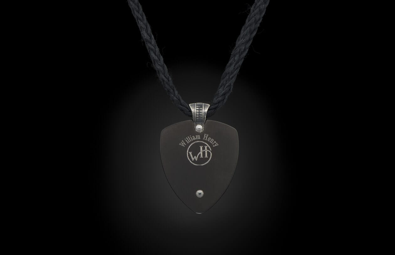 A dark grey pendant by William Henry, shield-shaped, with a silver rivet and etched logo, on a black braided cord.