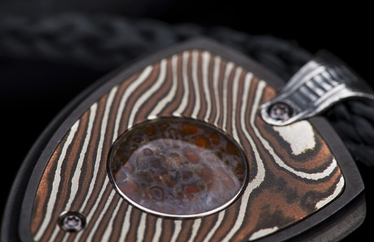 Decorative jewelry pendant featuring patterned copper and silver metal with a central mottled reddish-brown stone and ribbed silver bail on a black cord.