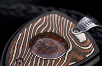 Decorative jewelry pendant featuring patterned copper and silver metal with a central mottled reddish-brown stone and ribbed silver bail on a black cord.