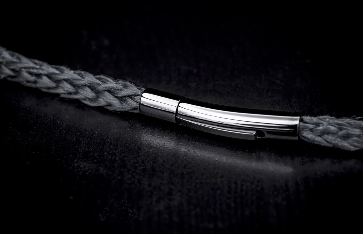 A dark grey braided bracelet with a sleek, polished silver-tone clasp on a dark textured surface.