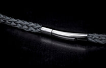 A dark grey braided bracelet with a sleek, polished silver-tone clasp on a dark textured surface.