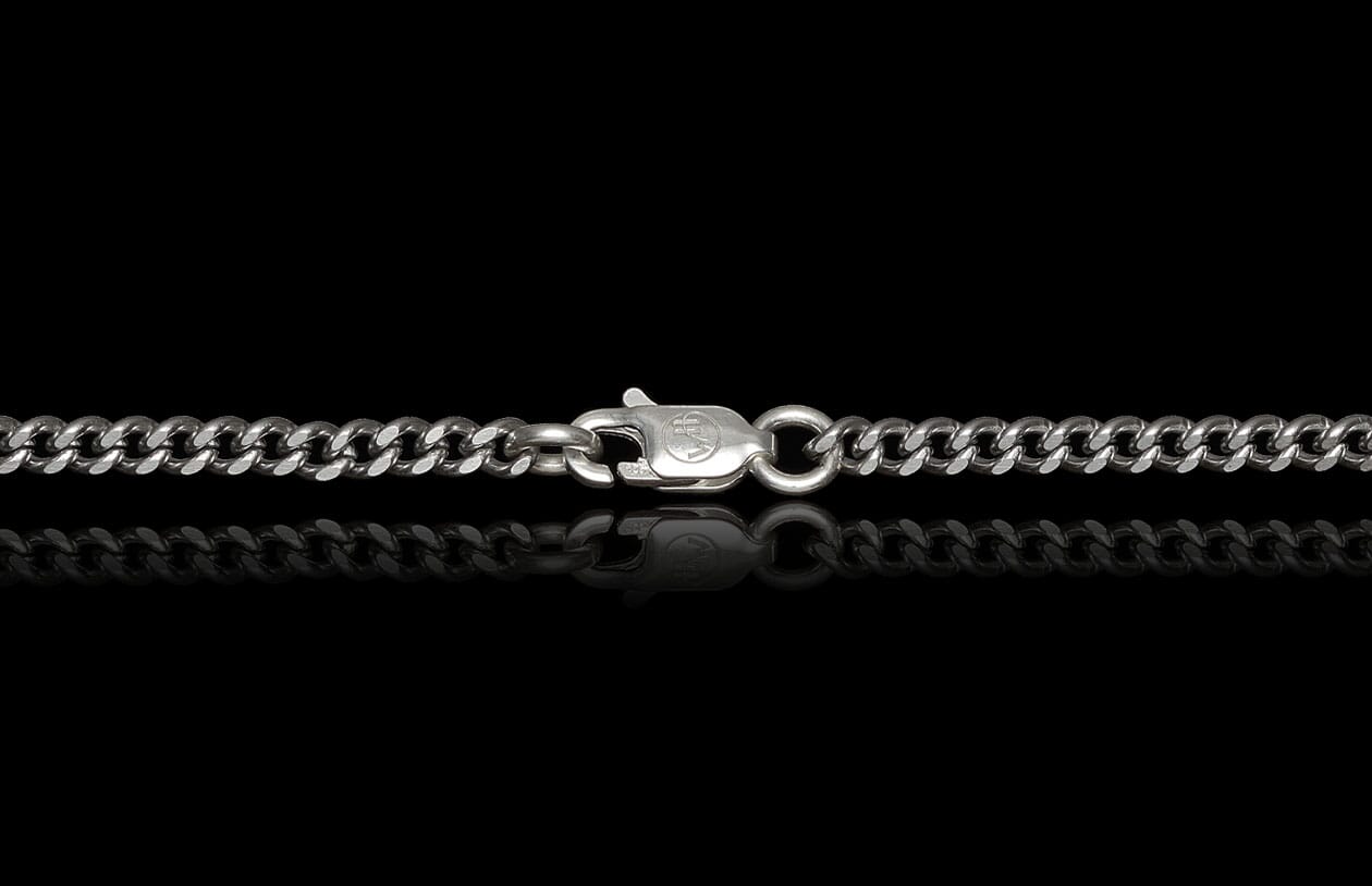 Silver curb chain by Mokume Forge with a lobster clasp featuring a circular emblem, reflected on black.