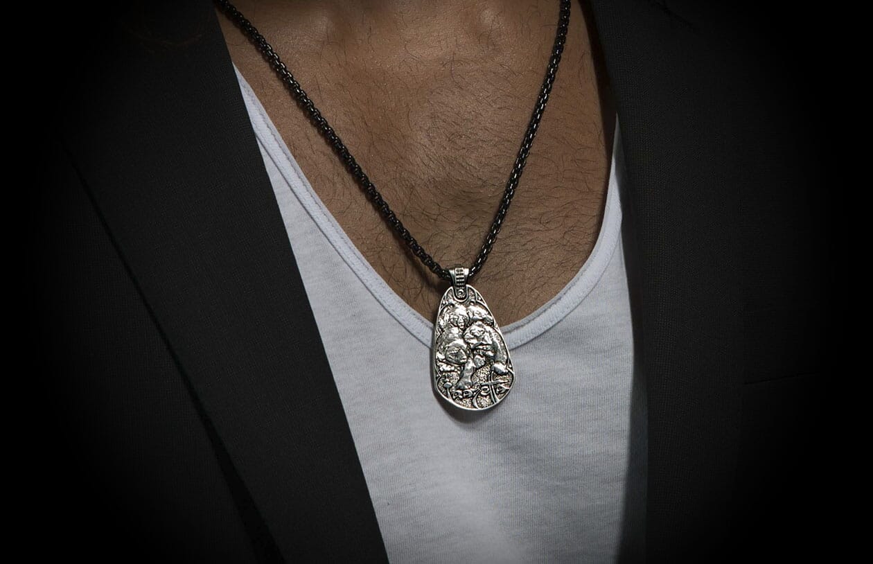 Silver Guardians pendant necklace with detailed relief carving, on a dark textured chain, worn over a white undershirt and black blazer.