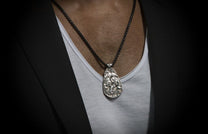 Silver Guardians pendant necklace with detailed relief carving, on a dark textured chain, worn over a white undershirt and black blazer.