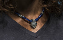 Sodalite shield necklace with blue beaded stones and a silver-toned shield pendant, worn on a person's neck.