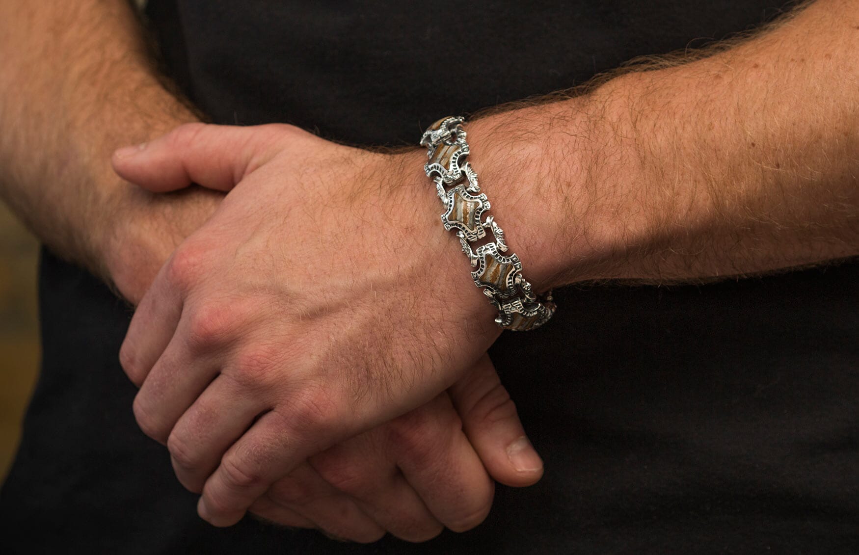 Ornate silver-toned bracelet with tan segments and intricate metalwork, worn on a man's wrist with hands clasped.