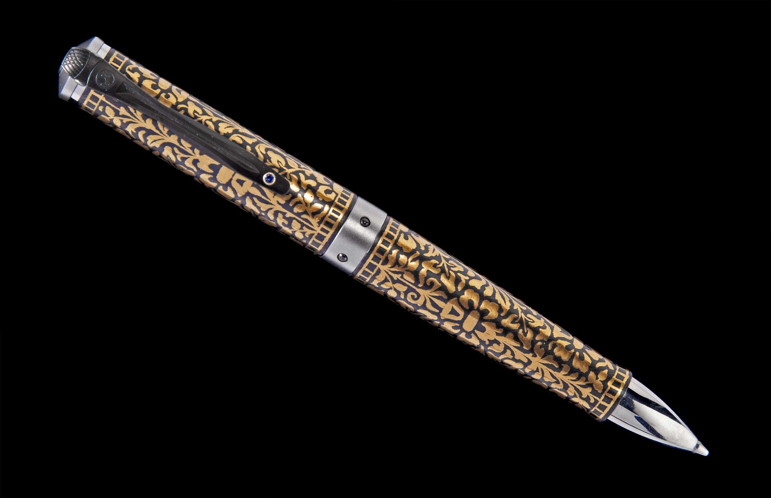 Black and gold pen with intricate floral pattern, silver accents, and a blue gem on the clip, angled on a black background.