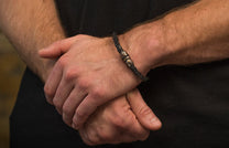 Black braided leather bracelet with a textured metal clasp and a small yellow stone, worn on a man's wrist.